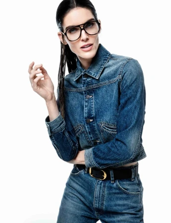 Portrait of an adult woman wearing denim jacket and jeans, accessorized with a belt and glasses, on a clean white backdrop.