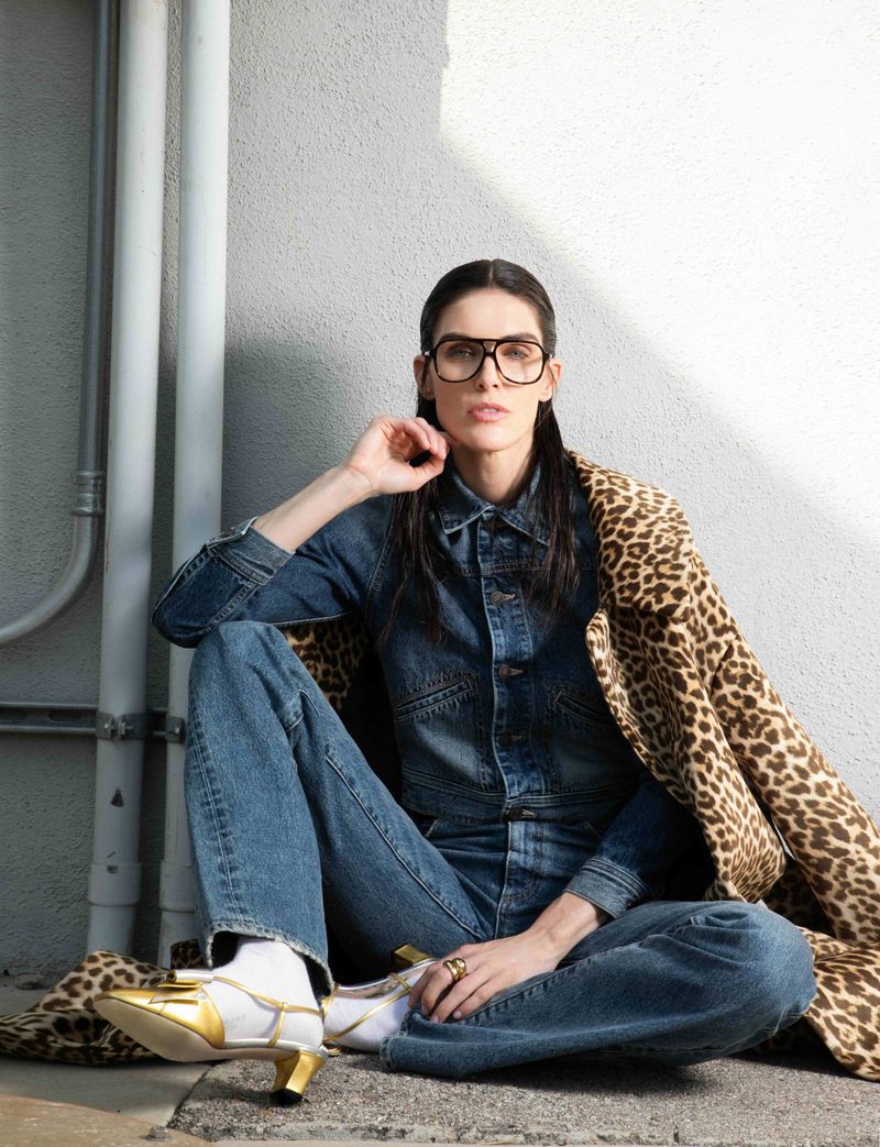 A woman with glasses and a denim outfit sits outdoors with gold shoes and a leopard-print coat, next to white pipes against a concrete wall.