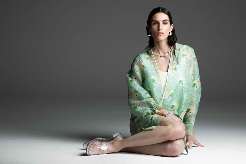 Elegant woman in floral jacket sits in a photo studio, showcasing transparent heels, accessorized with pearl necklace and earrings.