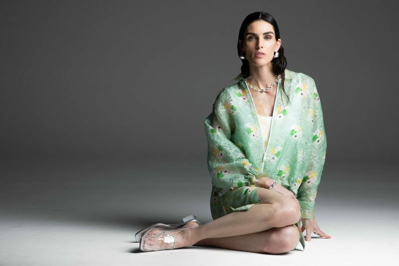 Elegant woman in floral jacket sits in a photo studio, showcasing transparent heels, accessorized with pearl necklace and earrings.