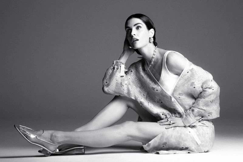 Black and white portrait of a young woman in a patterned jacket, tank top, statement jewelry, and clear heels posing indoors on a gray backdrop.
