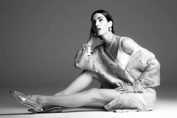 Black and white portrait of a young woman in a patterned jacket, tank top, statement jewelry, and clear heels posing indoors on a gray backdrop.