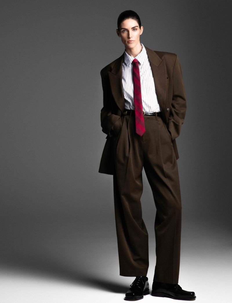 A person in a brown suit and red tie stands in a full shot against a grey backdrop in a fashion studio, exuding a confident pose.
