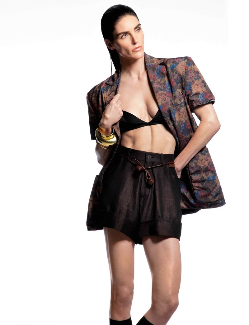An adult female poses in a photo studio wearing a black bra, brown shorts, and a patterned short-sleeved blazer against a white backdrop.