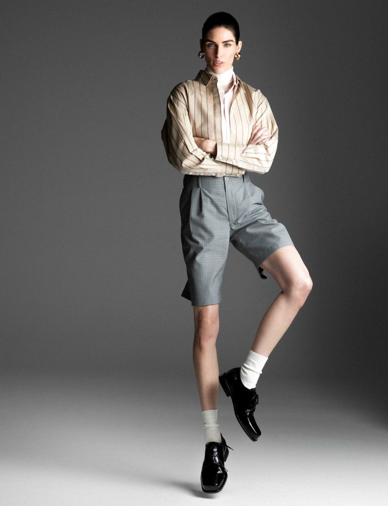 Woman in a striped shirt and grey shorts stands balancing on one leg in shiny black shoes and white socks against a grey background.