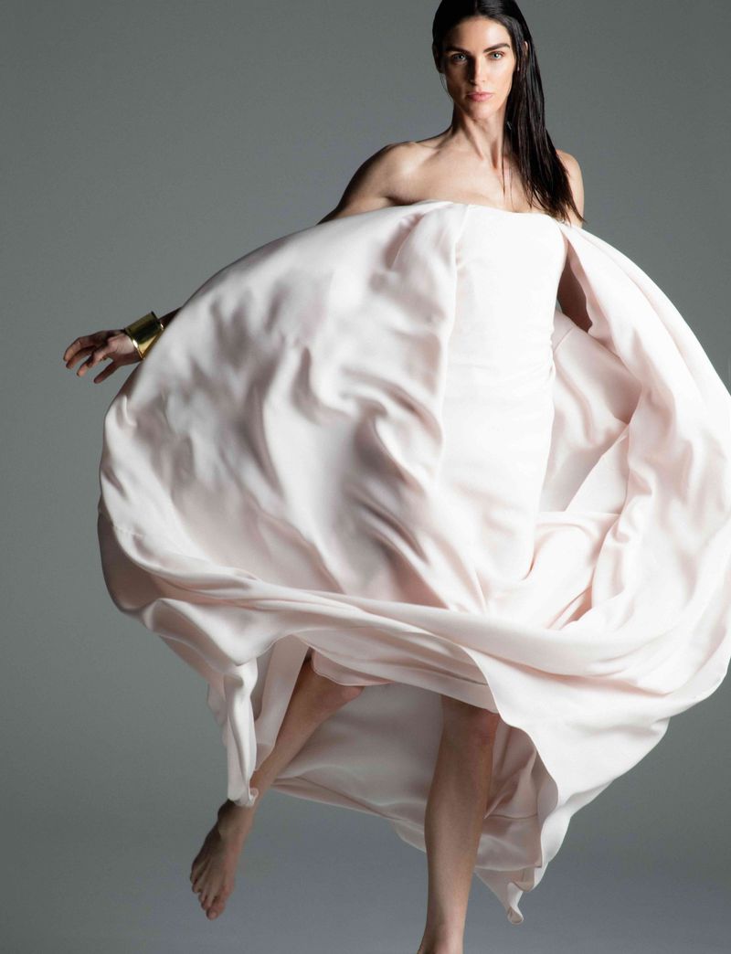 Fashion portrait of an adult woman in a flowing pale pink gown, accessorized with a gold cuff bracelet, against a neutral grey backdrop.
