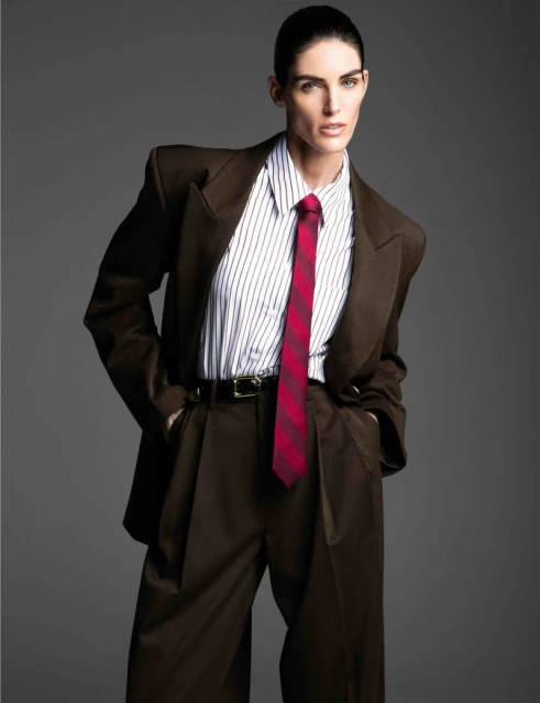 Fashionable woman in a brown oversized suit with a striped shirt and red tie posing in a studio with a neutral gray backdrop.
