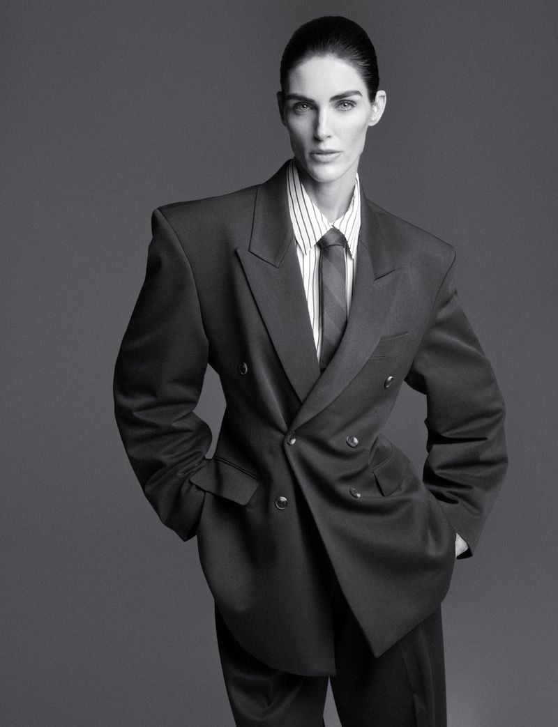 Monochrome studio shot of a person in a striped shirt and oversized suit jacket, posing with hands in pockets against a gray background.