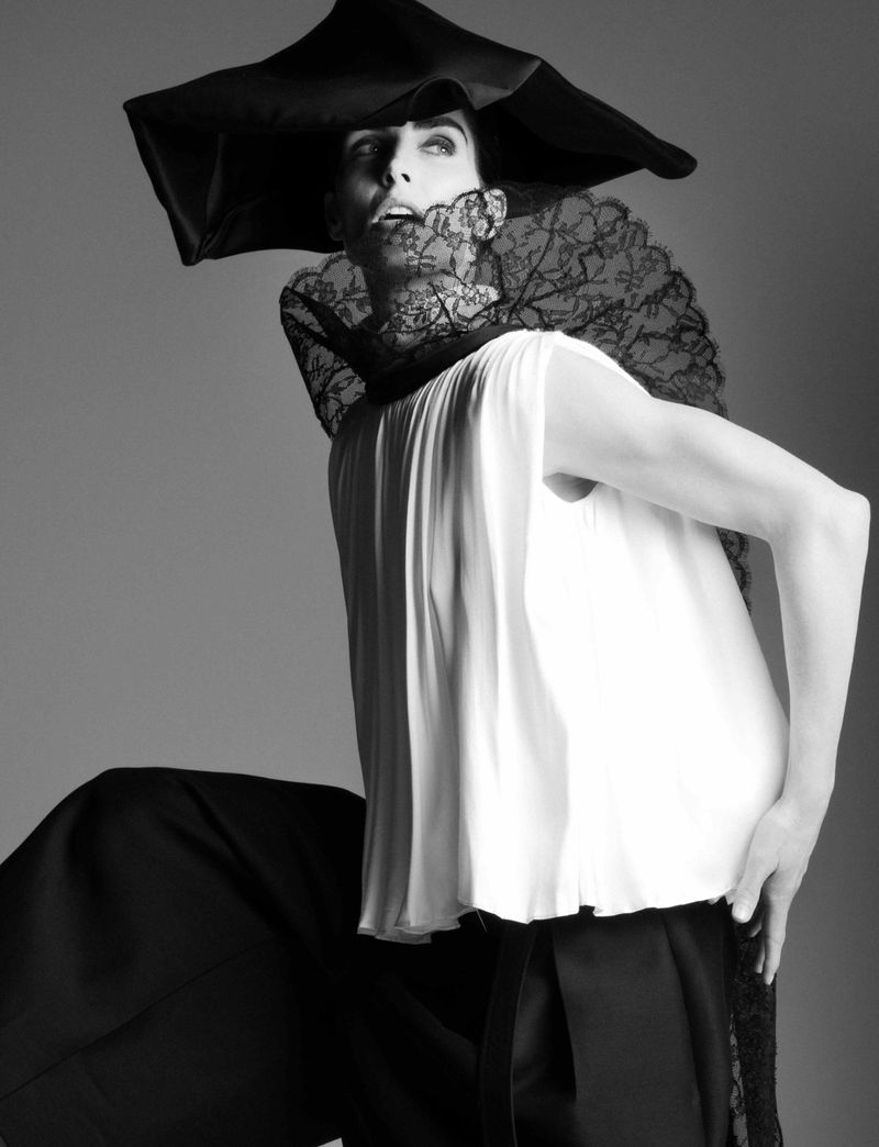 A person wearing a large black hat and lace collar poses in a black and white fashion photograph with a solid gray background.