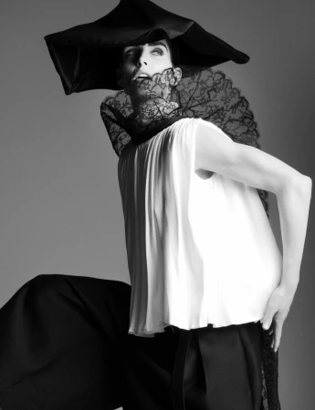 A person wearing a large black hat and lace collar poses in a black and white fashion photograph with a solid gray background.