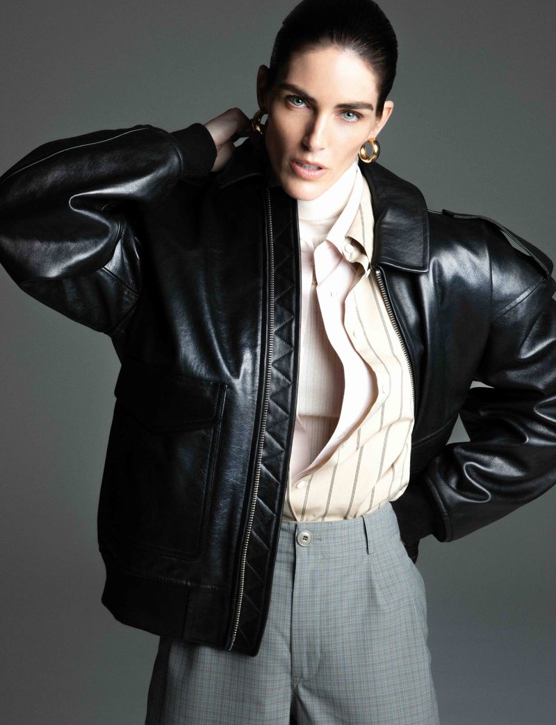 Portrait of a stylish adult woman in a black leather jacket, striped shirt, and gray plaid pants against a neutral gray backdrop.