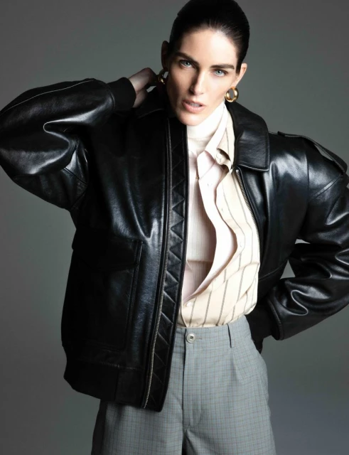 Portrait of a stylish adult woman in a black leather jacket, striped shirt, and gray plaid pants against a neutral gray backdrop.