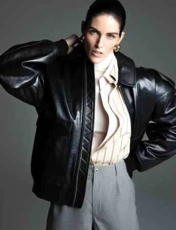 Portrait of a stylish adult woman in a black leather jacket, striped shirt, and gray plaid pants against a neutral gray backdrop.