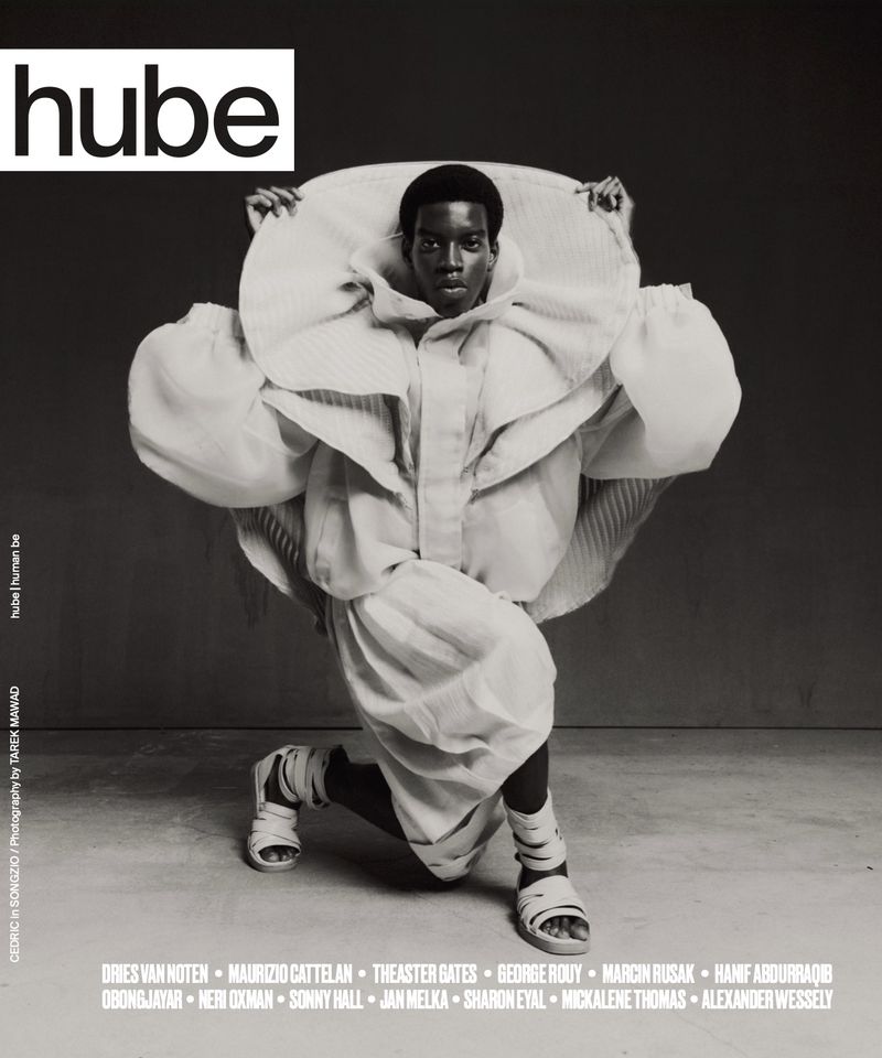 An African American model poses in an oversized white dress with a large collar, wearing strappy sandals in a studio shot for Hube.