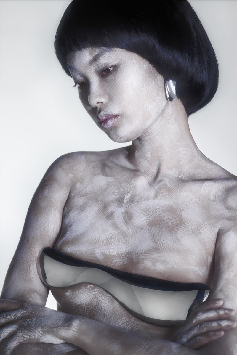 Elegant Asian woman with short black hair and silver body paint poses indoors, showcasing shoulder and chest with folded arms.