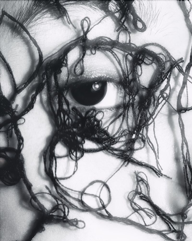 Close-up monochrome photo of a human eye surrounded by tangled dark thread or yarn, creating an abstract and textured composition.