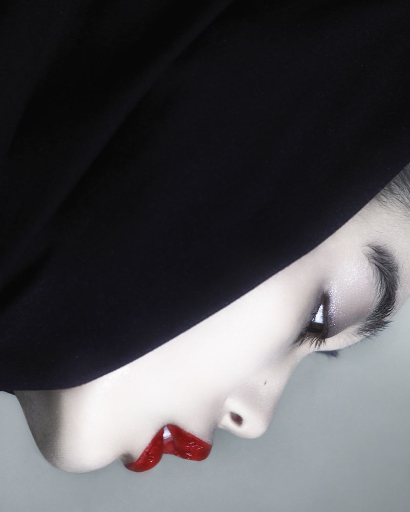 Close-up studio shot of a woman's face in profile, featuring bright red lipstick, dramatic eye makeup, and a black head covering.