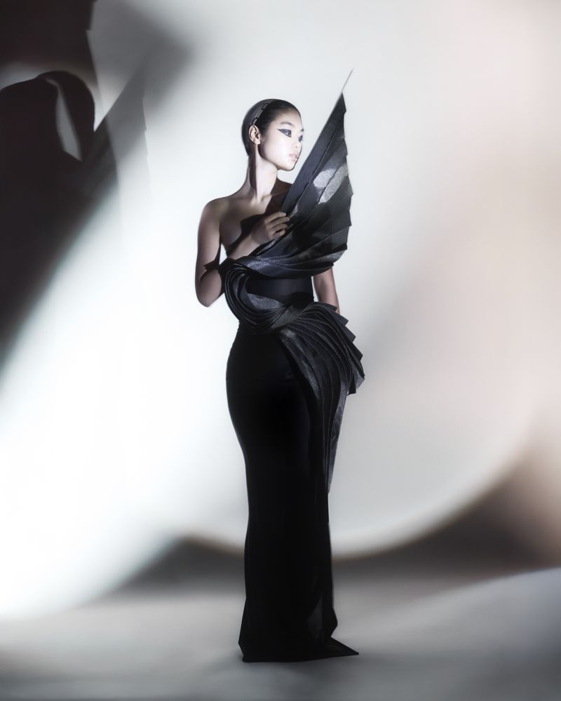 Elegant Asian woman in a black sculptural gown poses in a bright studio with soft light. Fashion editorial photography.