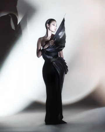 Elegant Asian woman in a black sculptural gown poses in a bright studio with soft light. Fashion editorial photography.