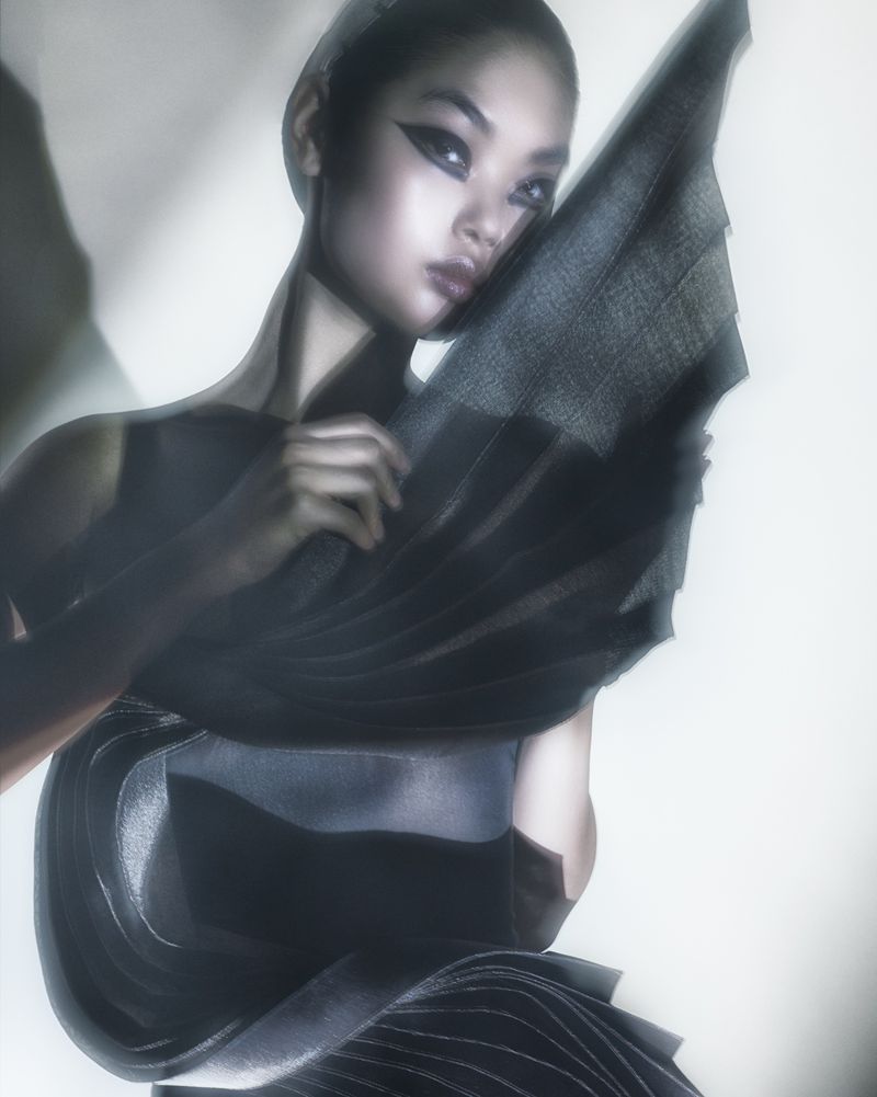 Striking fashion shot of an Asian woman with dramatic makeup, draped in elegant gray fabric, posing in a soft, diffused studio light.