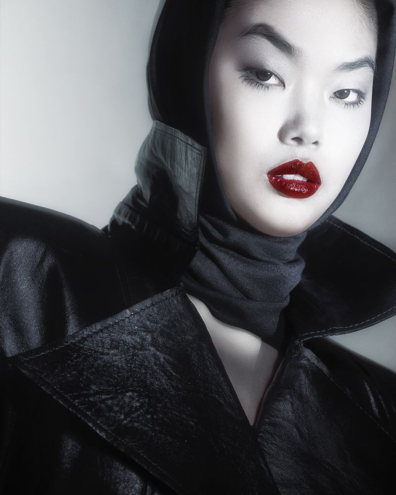 Glamorous close-up portrait of an Asian woman with red lipstick wearing a black leather coat and hood against a gray background.