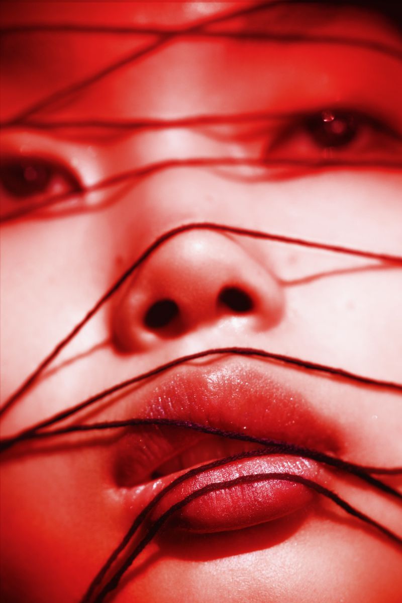 Close-up of a face with dark strings wrapped around the features in a red-toned image. The eyes, nose, and lips are visible.