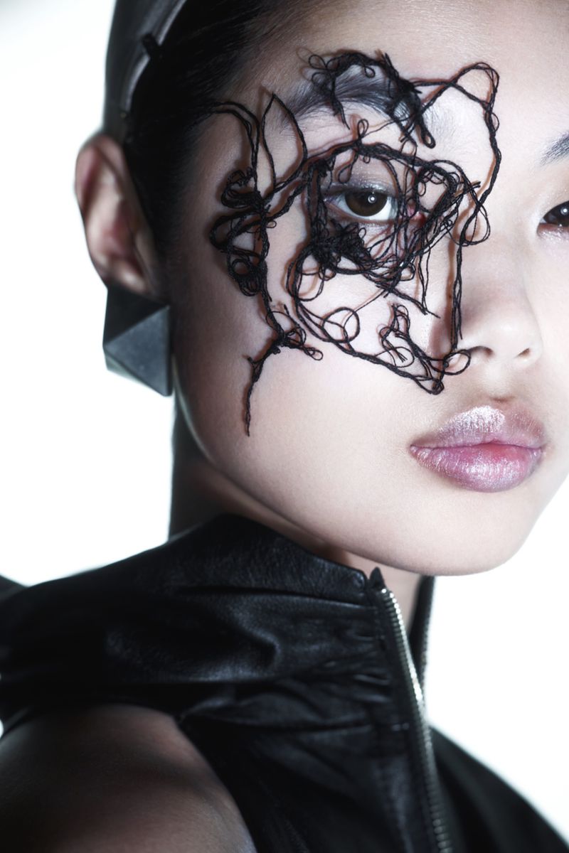 Asian woman with striking black thread eye makeup and leather jacket in a high-key fashion shot, highlighted by sharp geometric earring.