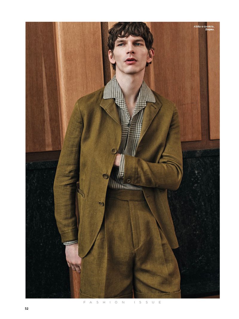 Man in an olive blazer and matching shorts stands against a wood and marble paneled wall in a fashion editorial shot.