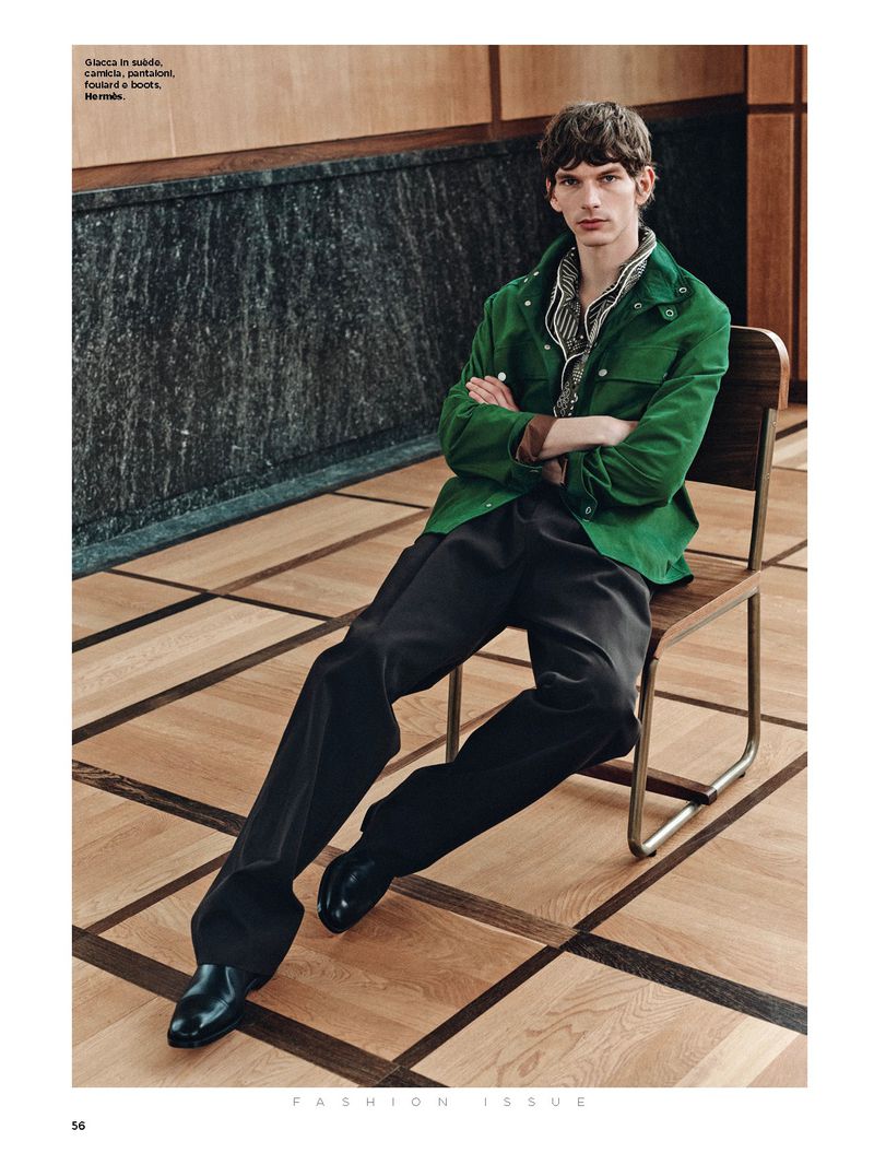 Man in a green jacket and black pants sitting on a wooden chair in an indoor setting with wood floors and a dark marble wall.