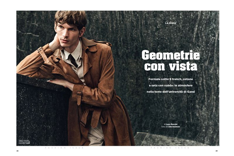 Editorial fashion shot of a young adult male model in a tan trench coat leaning against a stone ledge with a textured grey backdrop.