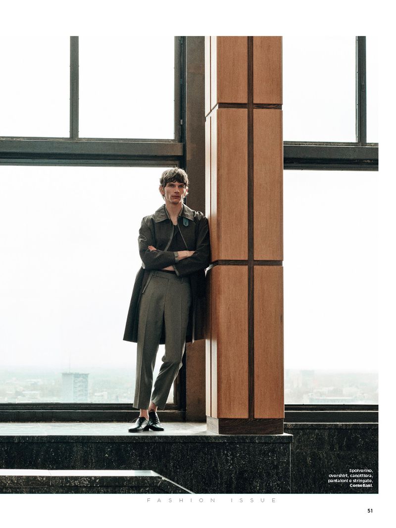 Fashionable man in coat poses indoors by large windows, leaning against an architectural column with muted city views. 