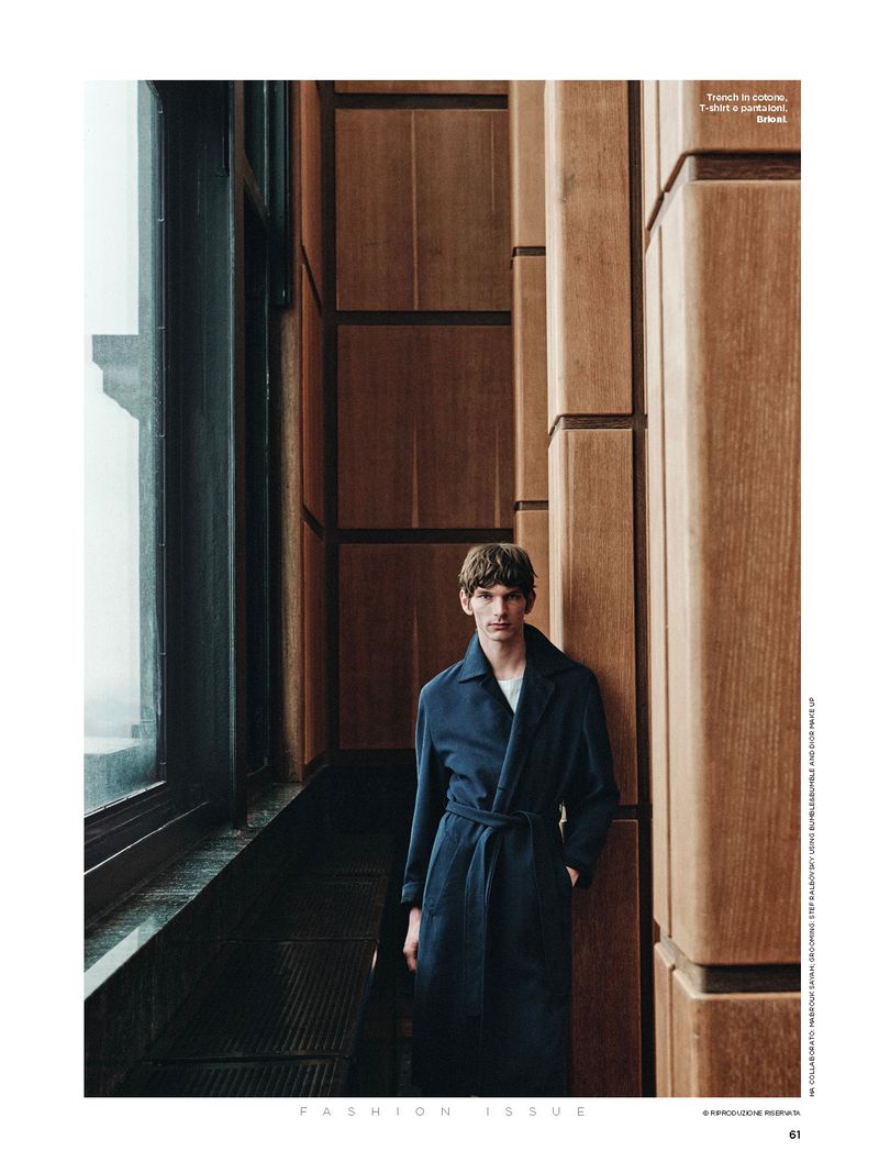 An adult male stands in front of a window and panelled wall wearing a dark blue belted coat in a fashion editorial shot.