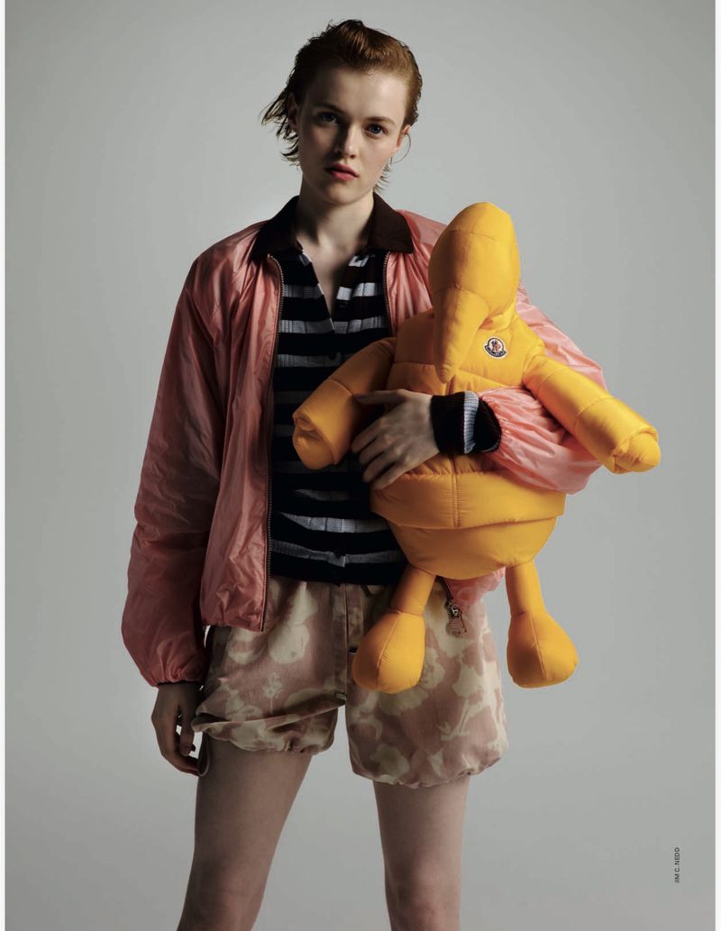 Woman in pink jacket holding yellow Moncler stuffed animal in a studio setting, showcasing fashion and editorial style with soft lighting.