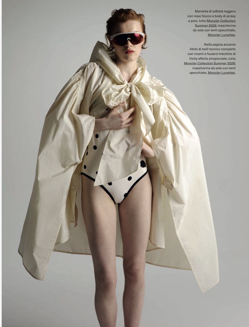 Fashion editorial shot of a redhead woman wearing a polka-dot swimsuit, sunglasses and oversized cream-colored coat with a bow.