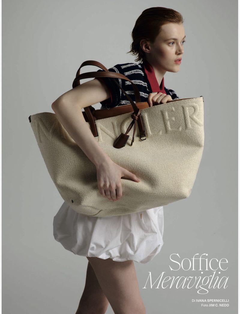 Fashion editorial shot of a stylish red-haired woman carrying a large beige fabric handbag with leather trim in a bright, neutral studio.