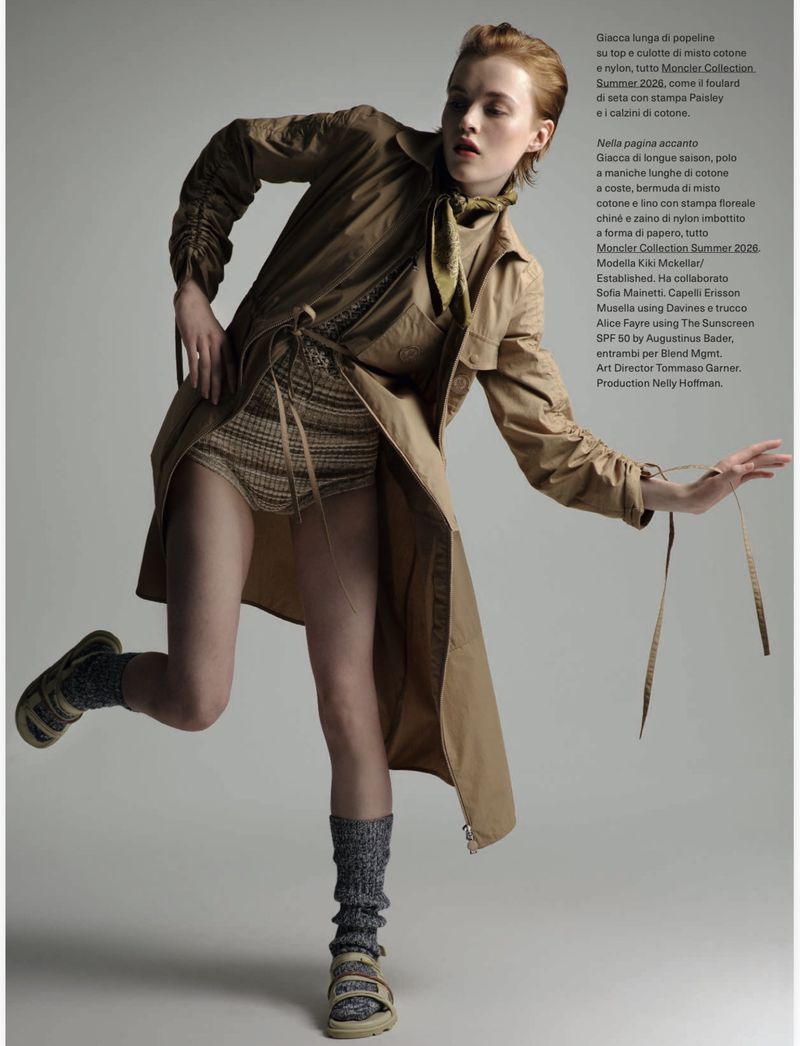 Fashion editorial shot of a woman in a tan trench coat, knit shorts, and sandals posing in a photo studio with a neutral background.