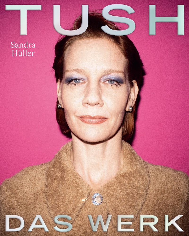 Close-up portrait of Sandra Hüller on TUSH magazine cover with silver metallic fonts against a pink background, and wearing a brown coat.