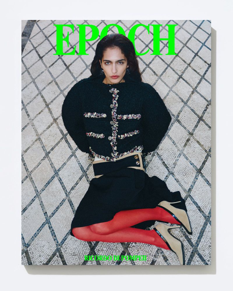 Magazine cover featuring a woman in a black jacket and skirt lying on a mosaic tile floor, with bold green text reading 'EPOCH'.