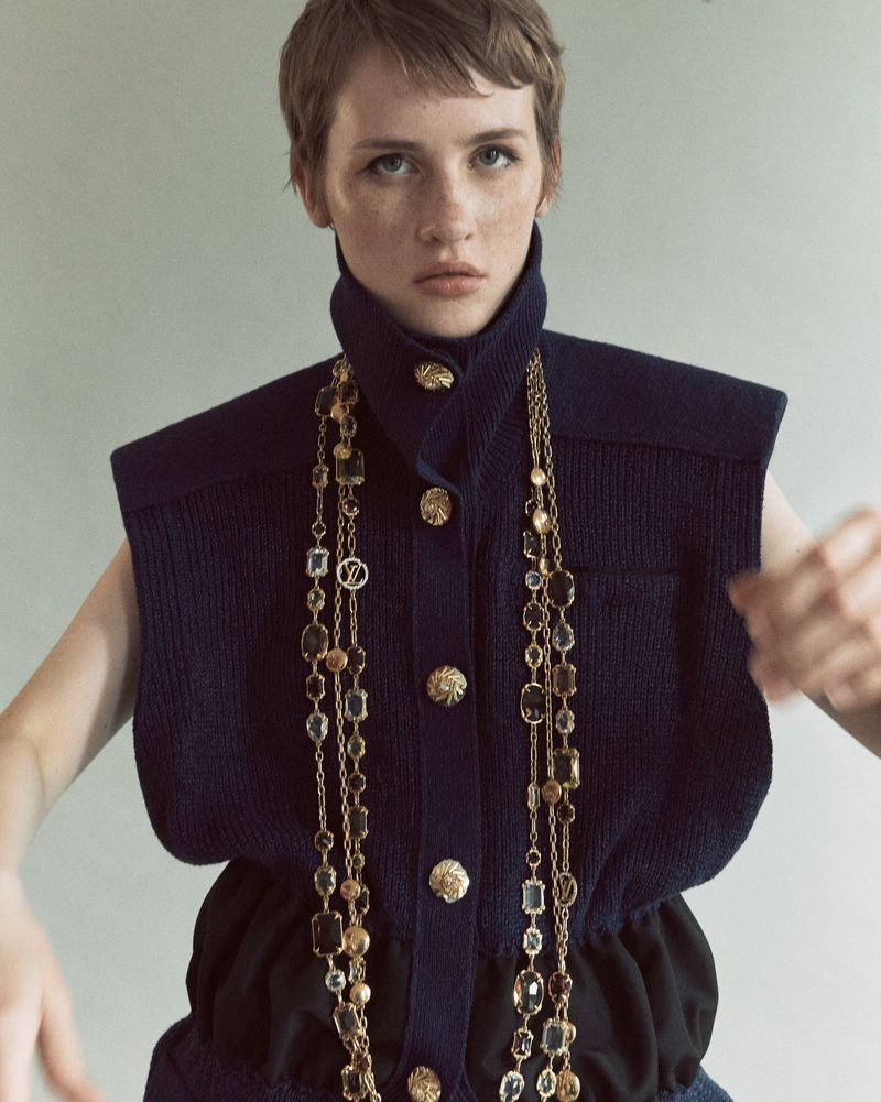 A fashion portrait of an adult female model with short hair wearing a sleeveless top and long chain necklace, posing indoors.