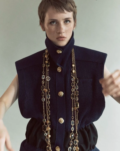 A fashion portrait of an adult female model with short hair wearing a sleeveless top and long chain necklace, posing indoors.