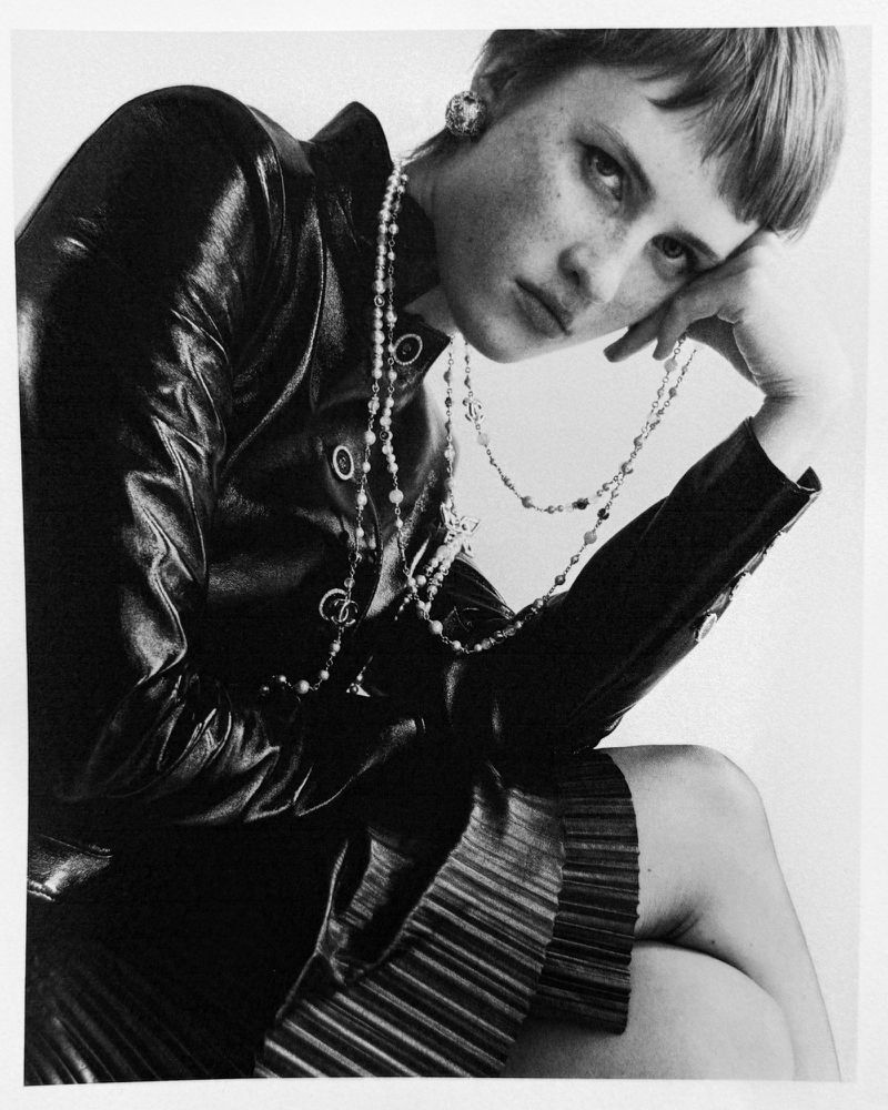 Black and white fashion shot of a stylish woman with short hair wearing a leather jacket and pearl necklaces in a studio setting.