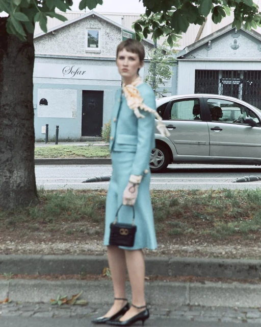 Stylish woman in light blue suit and heels with a small handbag, standing on a city street with a car and building in the background.