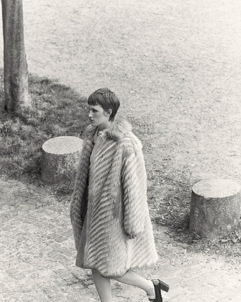 Monochrome portrait of a woman outdoors wearing a patterned fur coat, standing on a paved surface with foliage in the background.