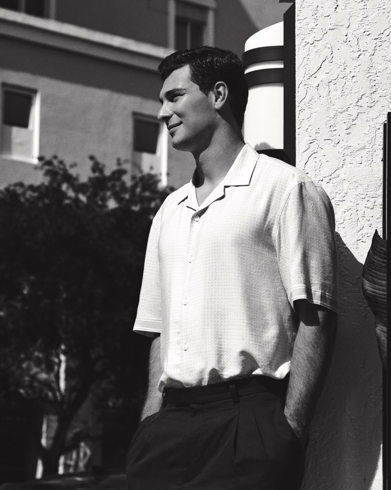 Monochrome portrait of a handsome man in a short-sleeve shirt and dark pants, leaning against a textured wall outdoors.