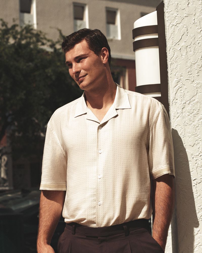 Adult male wearing an off-white button-down shirt and dark pants stands next to a stucco wall and a column with a light fixture.