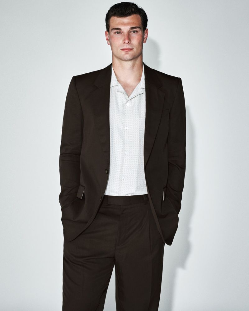 Man in brown suit with hands in pockets, standing indoors against a neutral backdrop, medium shot, professional studio lighting.