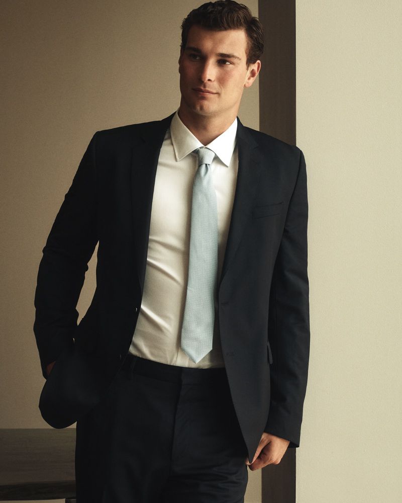 Handsome man in a black suit and blue tie posing indoors against a neutral wall. The shot is a medium shot with soft lighting.
