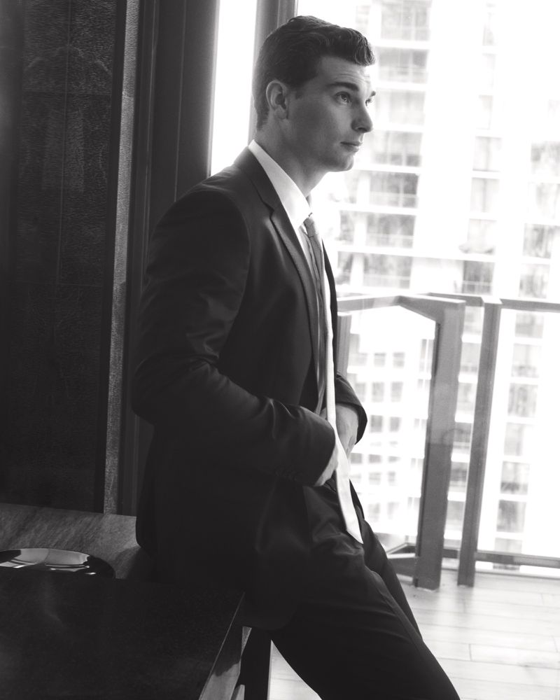 Monochrome portrait of a man in a suit looking out of a window, posing with a hand in his pocket in an indoor setting.