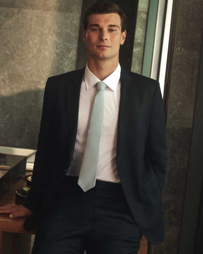 Professional headshot of an adult male in business attire, wearing a black suit and light blue tie, standing indoors.