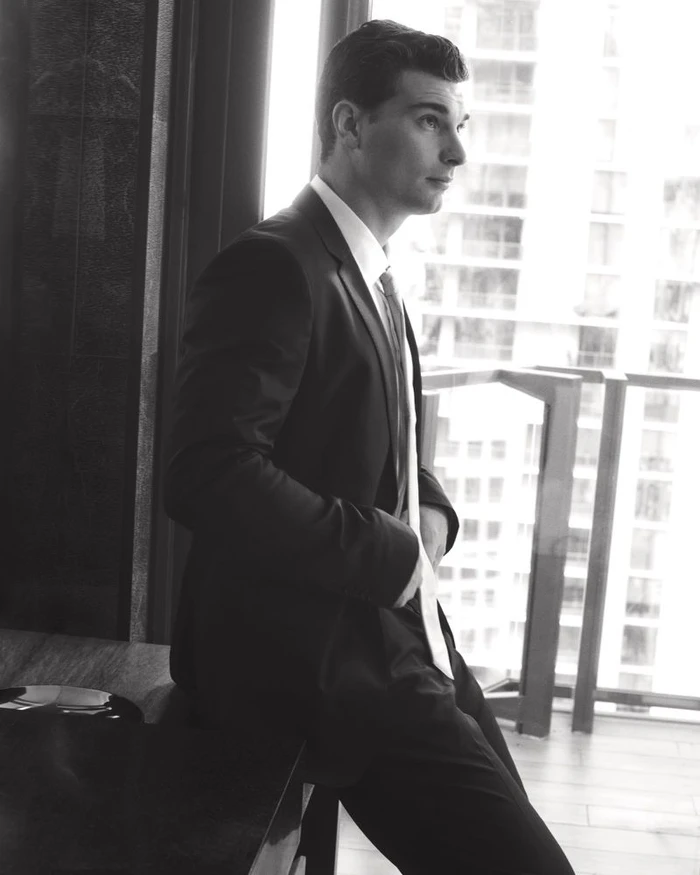 Black and white portrait of a man in a tailored suit leaning against a desk near a bright window.